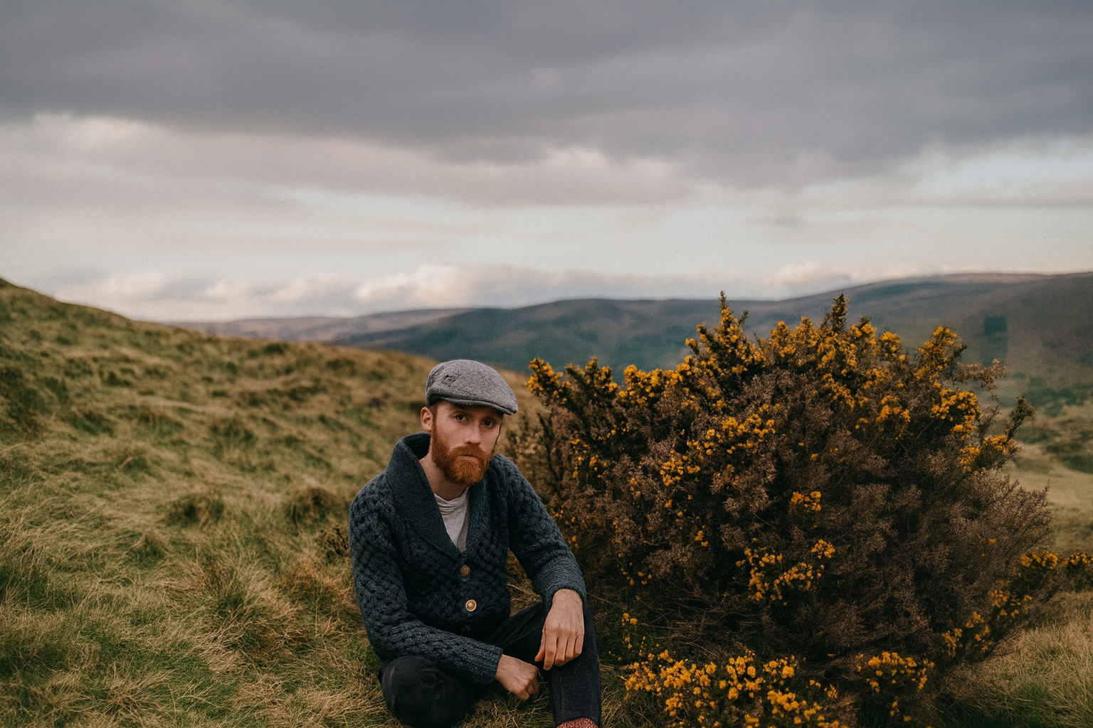 Man wearing shawl-collar Aran cardigan sitting beside yellow gorse on hillside.